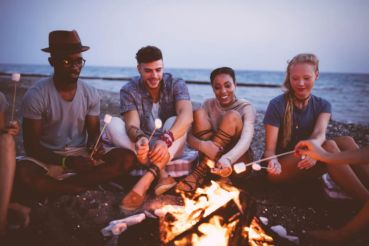 Happy friends roasting marshmallows over campfire at beach party