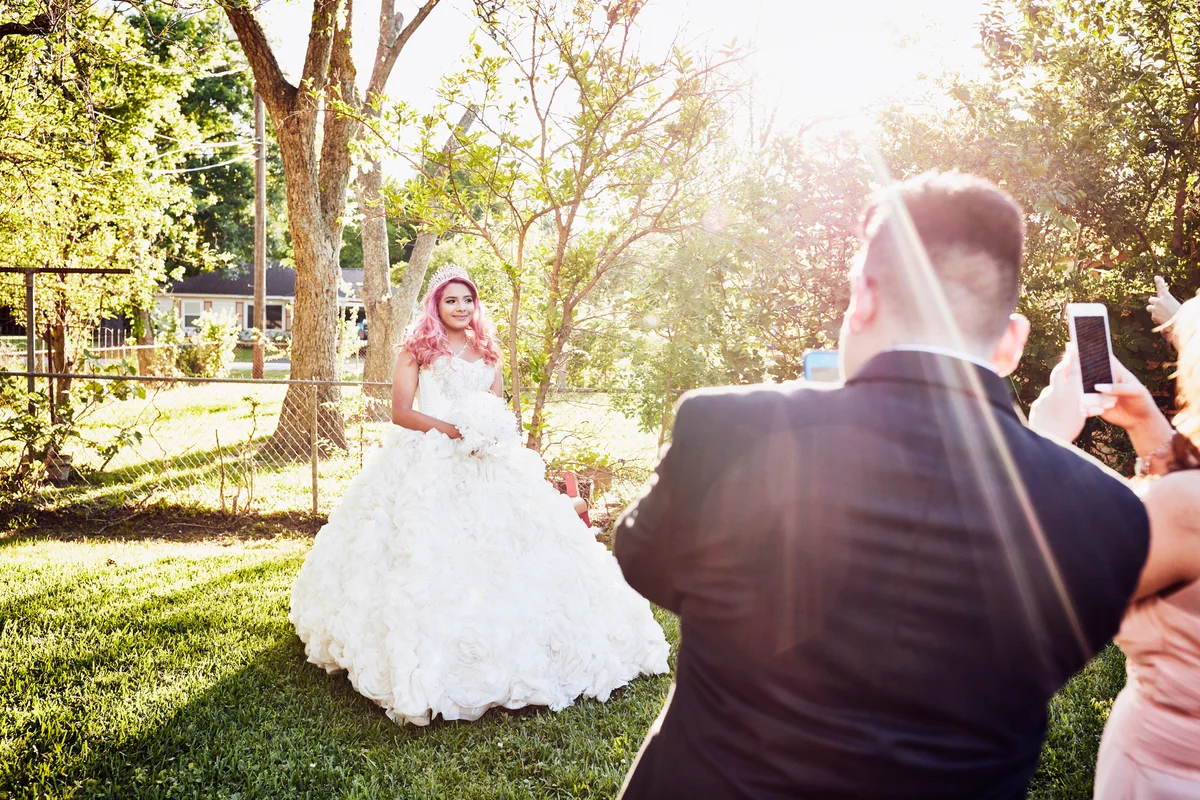 Family taking photos of young woman in quinceanera gown in backyard