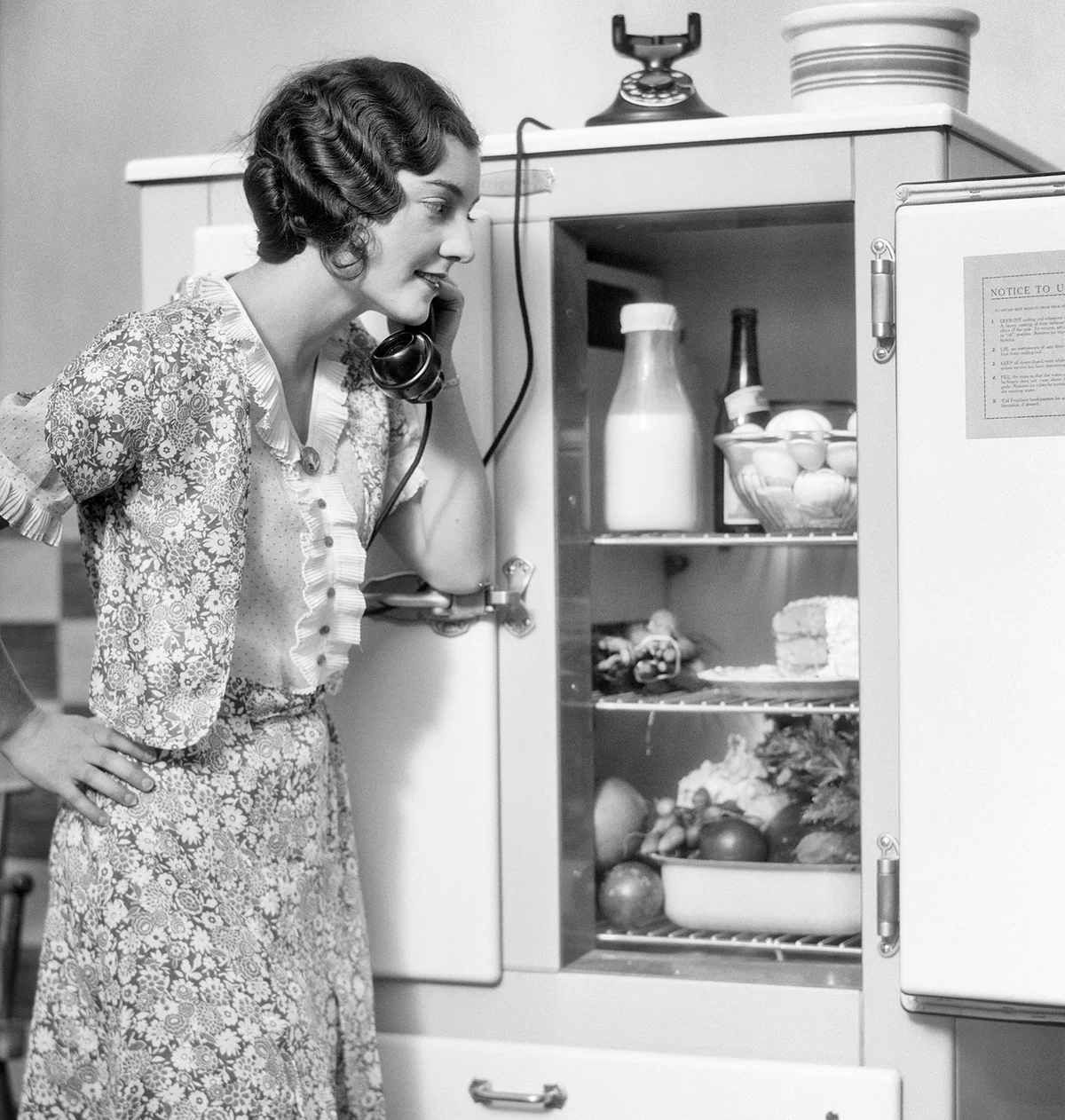 WOMAN SPEAKING ON TELEPHONE LOOKING INTO THE KITCHEN ICEBOX REFRIGERATOR - Getty Editorial Use