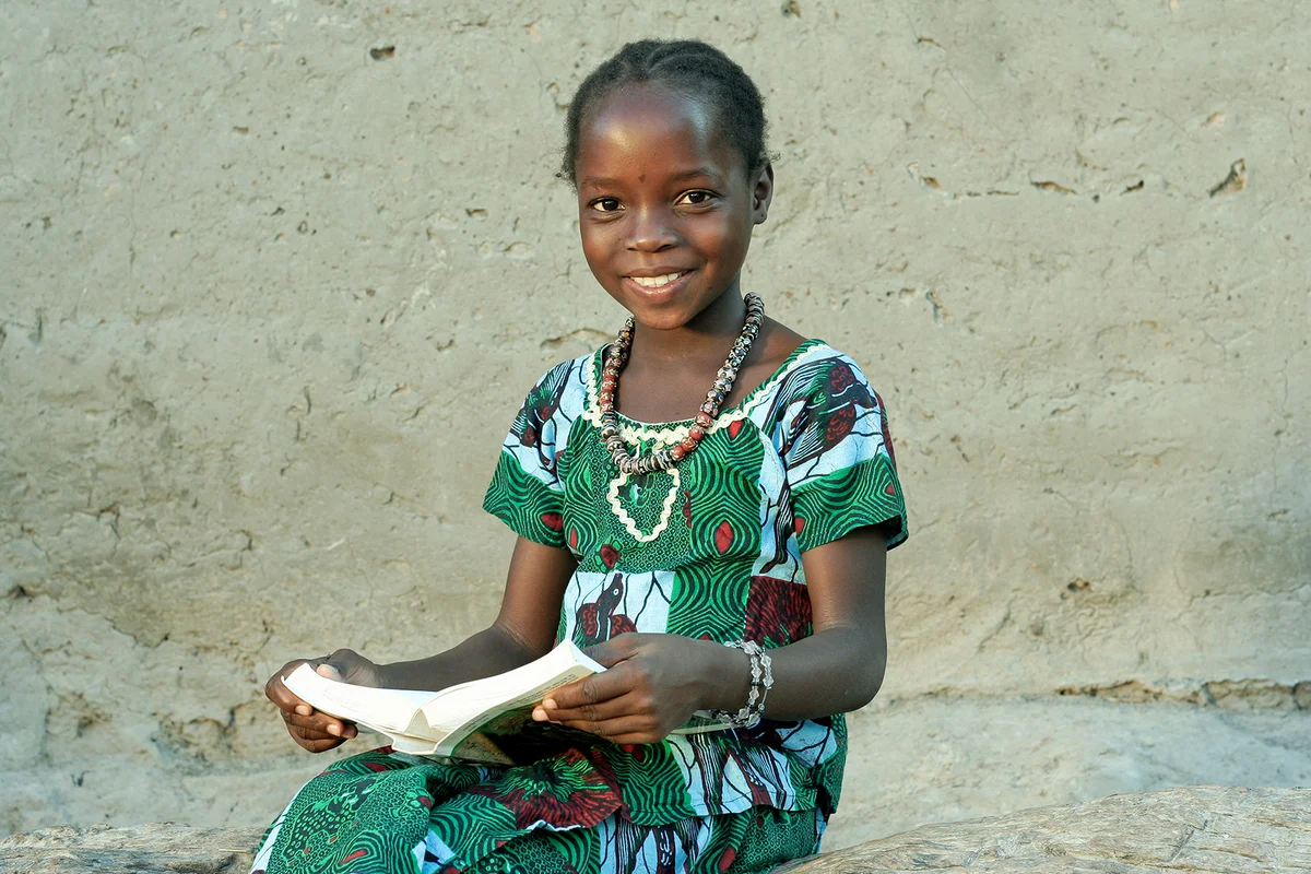 A young girl in a village, sitting with an open book on her legs, looking at the camera, smiling.
