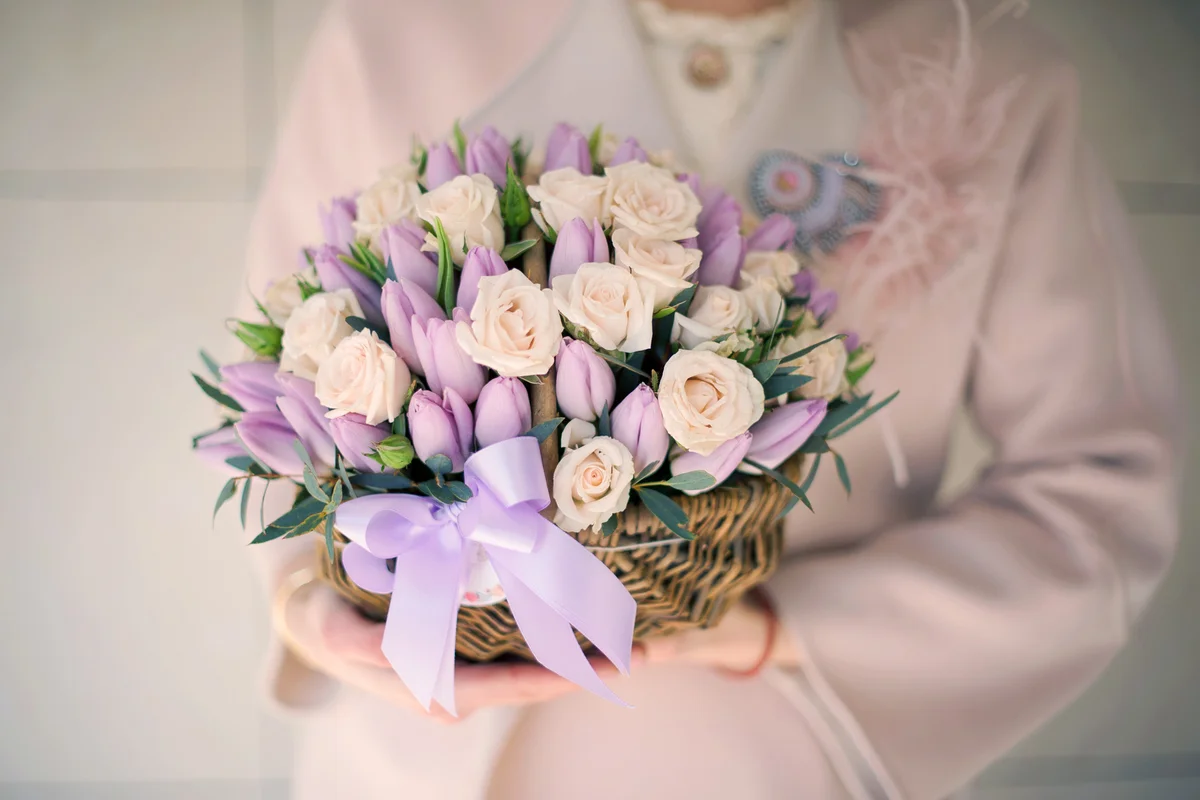 Girl in the coat holding a wicker basket of tender creamy color roses and violet tulips