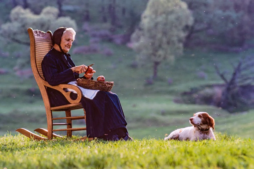 Elderly woman sits outdoors on a rocking chair peeling apples