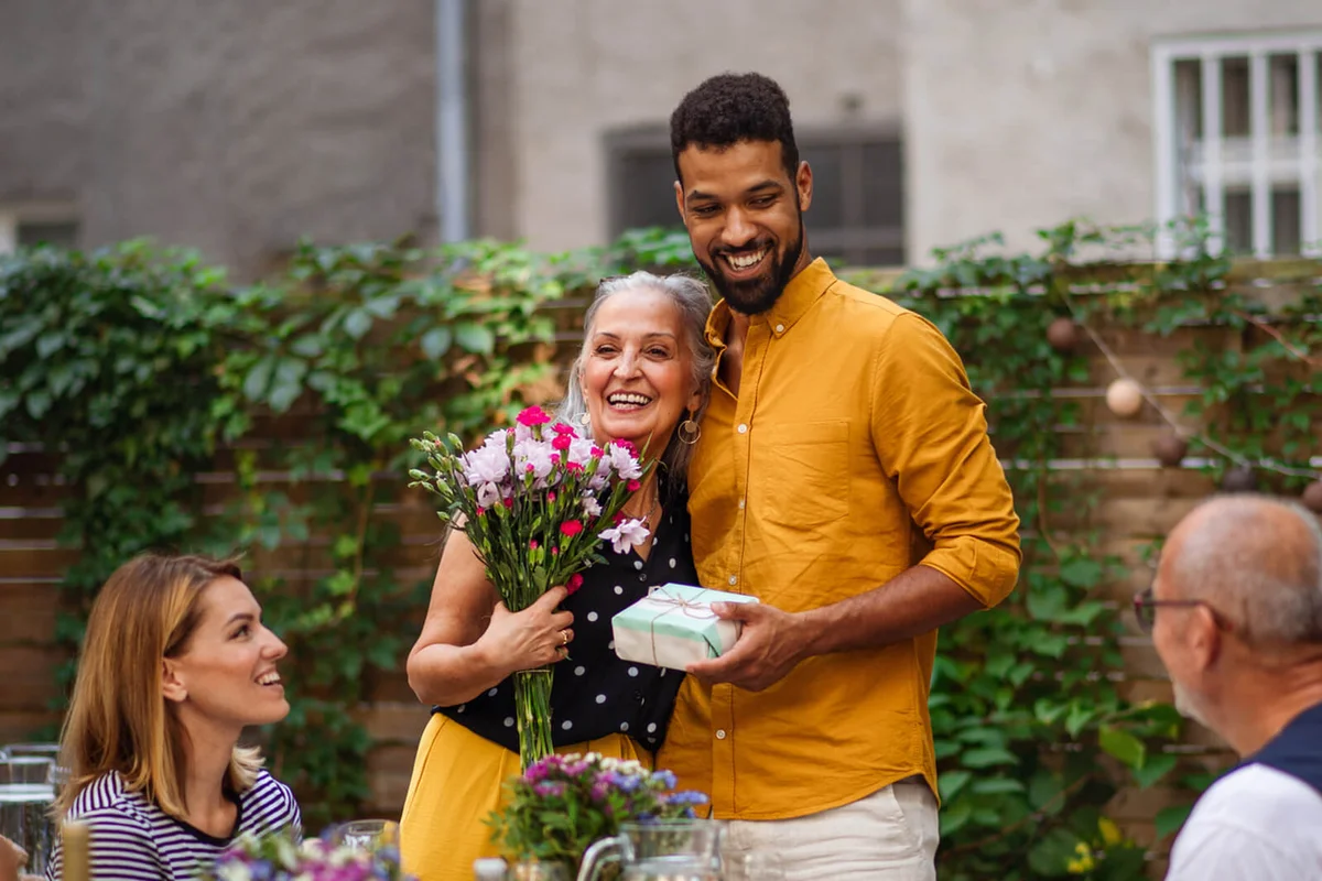Happy young man with bouquet and gift hugging his senior mother in law outdoors in garden