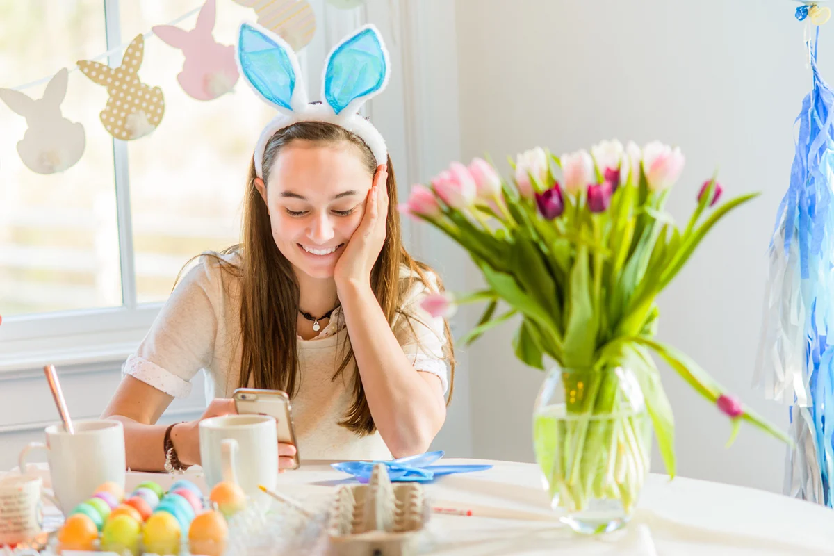 Teenage girl at table reading smartphone texts whilst dyeing hard boiled eggs for Easter