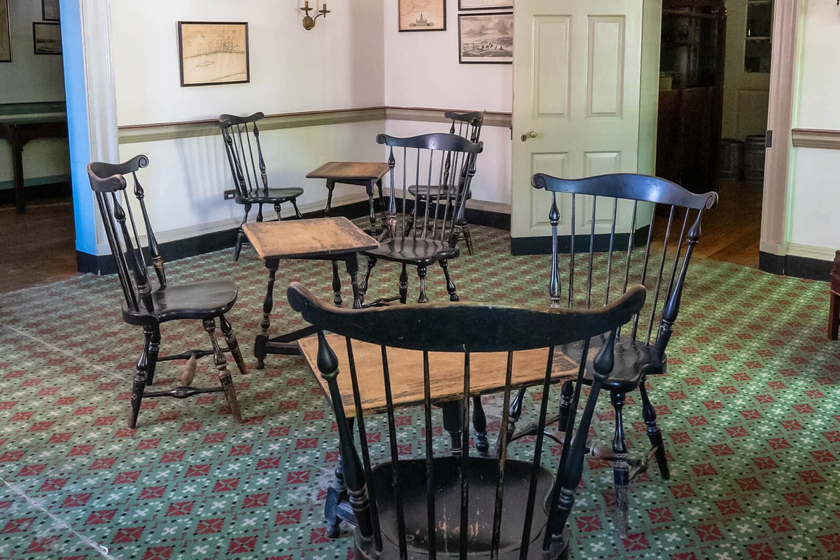 Room with groupings of Windsor chairs around tables all placed on a wall-to-wall red and green patterned carpet.