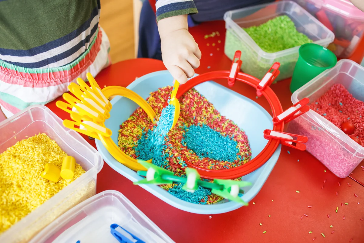 Toddlers playing with sensory bins with colorful rice on red table