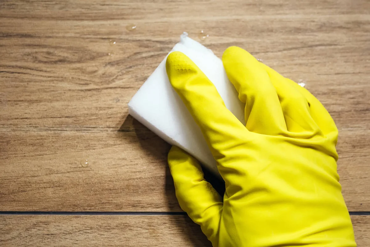 A hand in a yellow rubber glove wipes wet tile imitating a wooden surface with a white melamine sponge