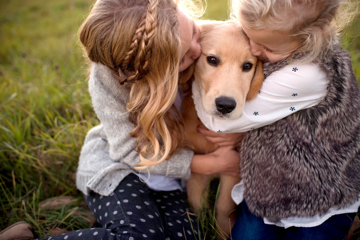 Two young girls outdoors hugging and kissing pet dog