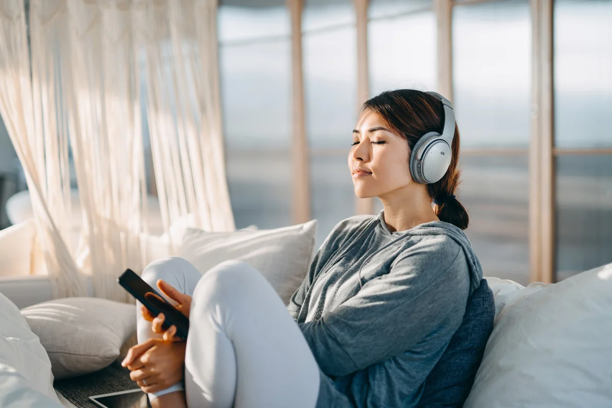 Relaxed young woman with eyes closed sitting on her bed enjoying music over headphones