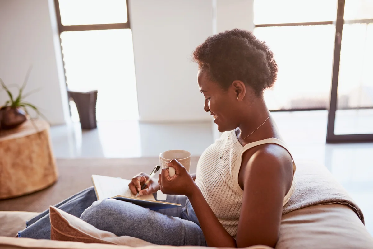 Young woman having tea and writing in a notebook on the sofa at home