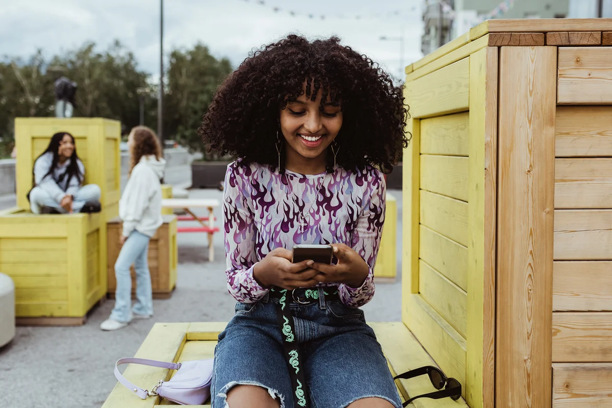 Smiling teenage girl using smart phone while sitting on box in city