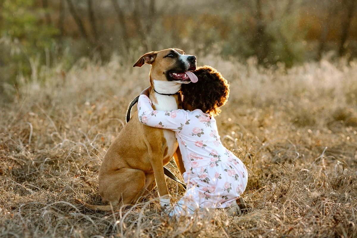 Side view of girl embracing dog while kneeling on grassy field at forest