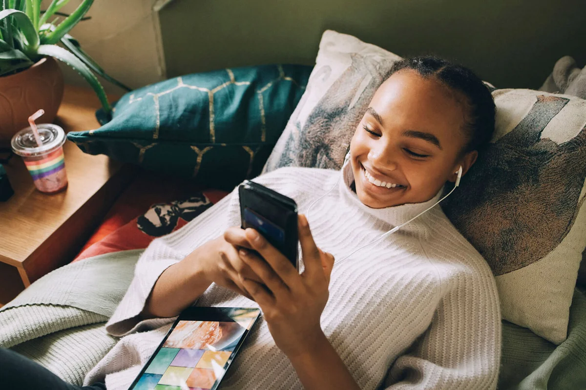 smiling teenage girl listening music while using mobile phone on bed at home