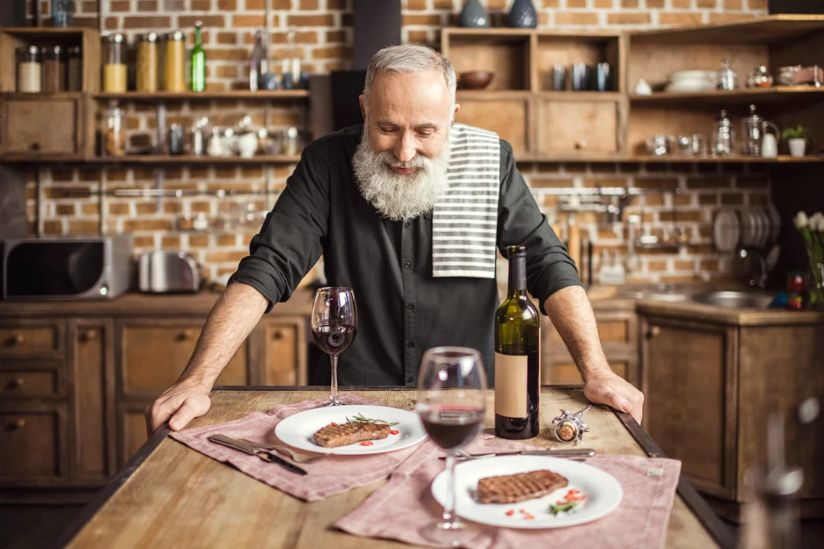 Smiling bearded man leaning at dining table with red wine and steaks