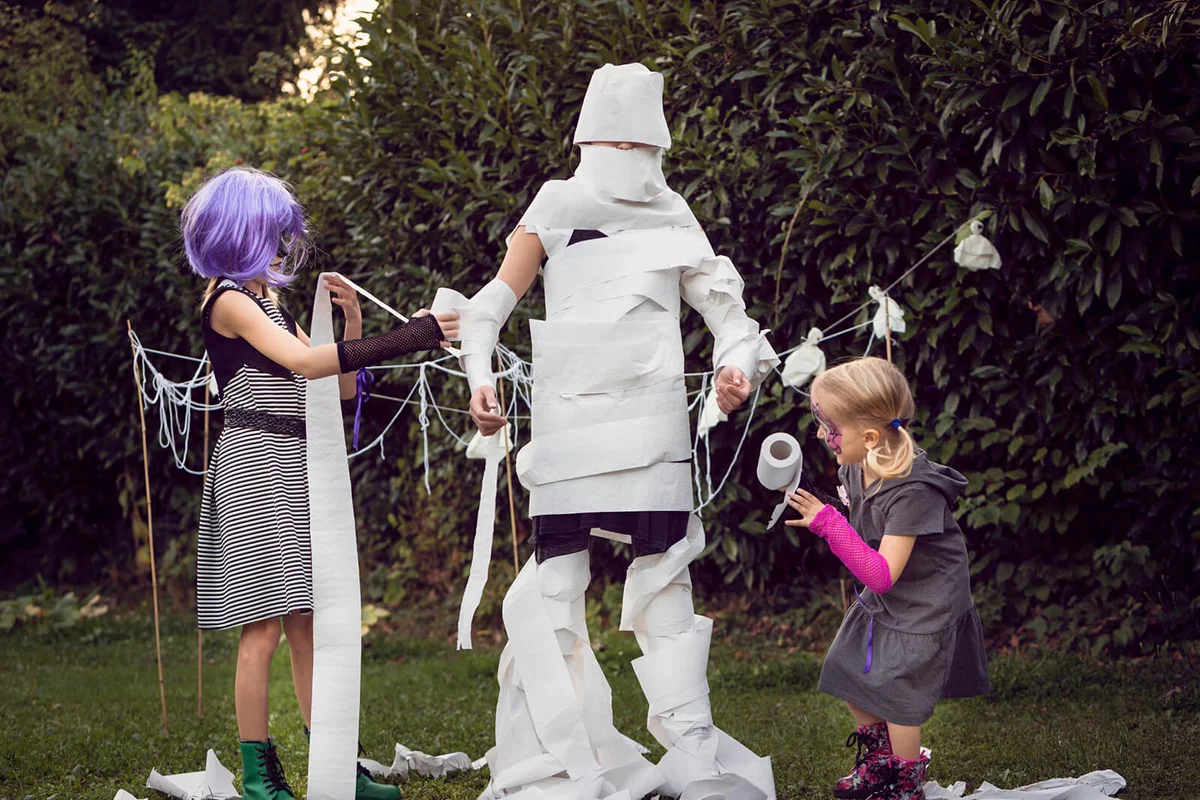 Two children wrapping a third child with toilet paper to play 'Mummy Wrap' Halloween game