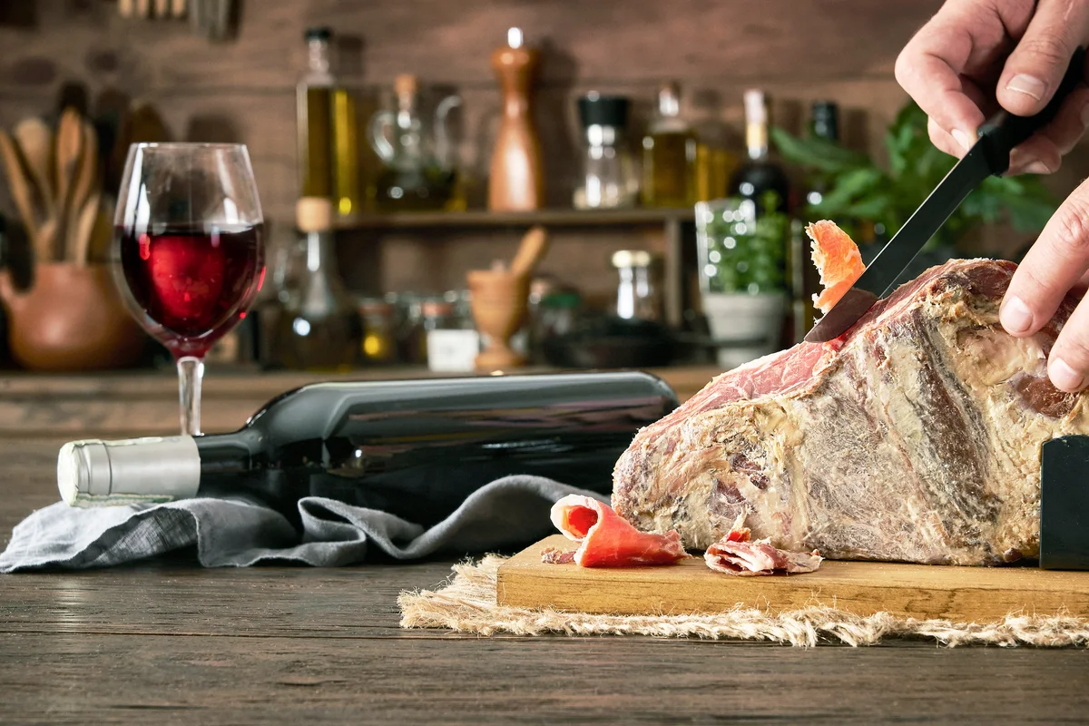 Man slicing Iberian pig ham leg on cutting board with bottle of red wine on rustic wooden table