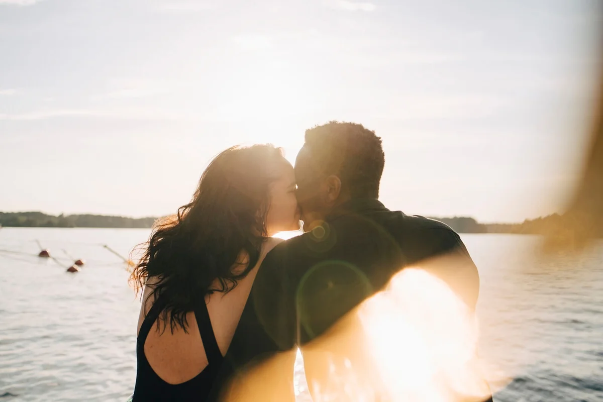 couple kissing while sitting against lake during summer
