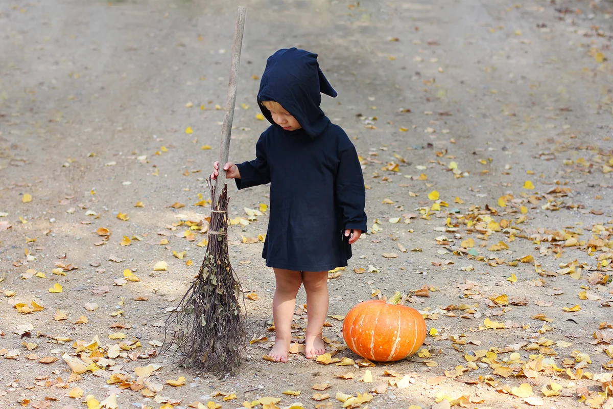 Child in black sorcerer or witch suit stands with broom near fresh pumpkin