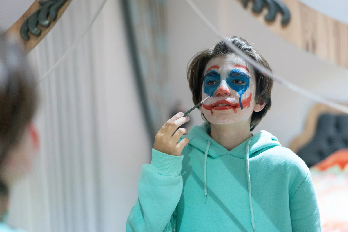 Young boy face painting for Halloween night party looking at mirror getting ready
