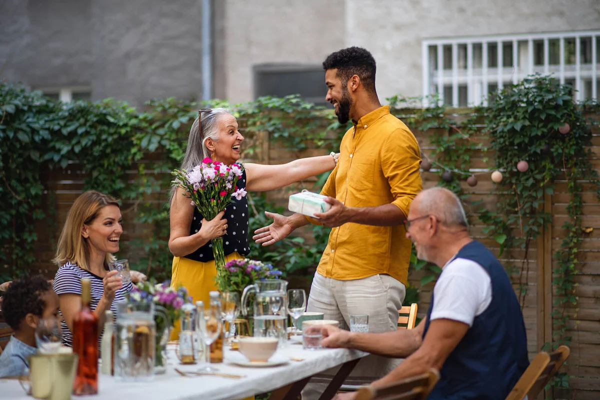 Happy young man with bouquet and gift congratulating his senior mother in law outdoors in garden