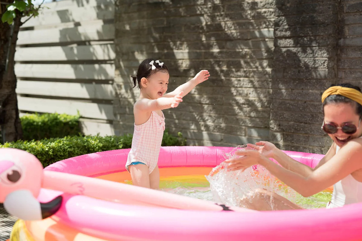 A toddler splashes water on her mother as both laugh while playing in an inflatable pink pool
