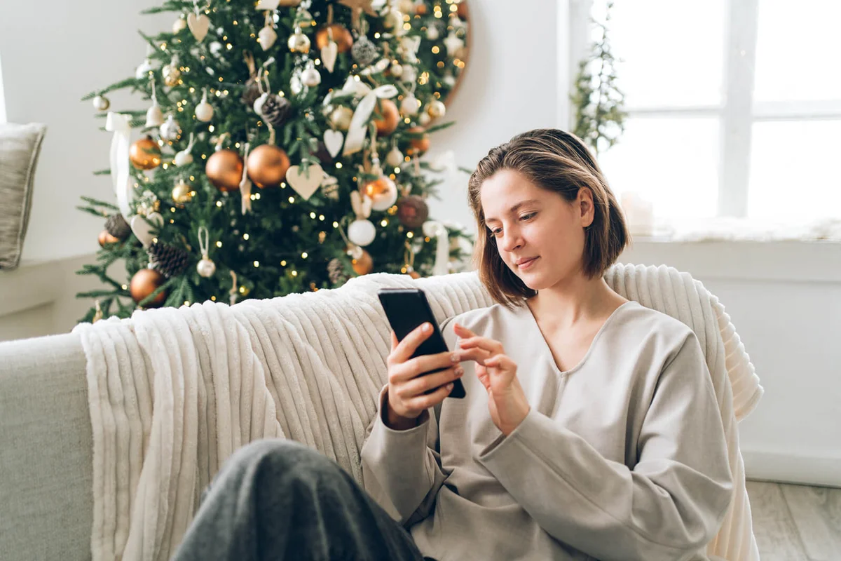Young woman is using smartphone sitting on sofa near decorated Christmas tree