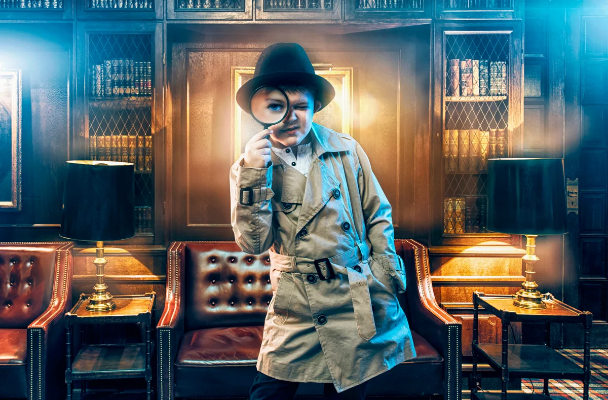 Boy detective wears a trench coat and a fedora hat using magnifying glass