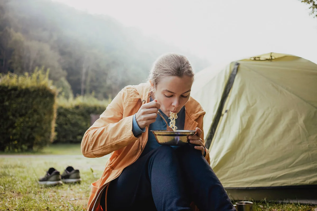 Female hiker eating noodles while sitting against tent in forest