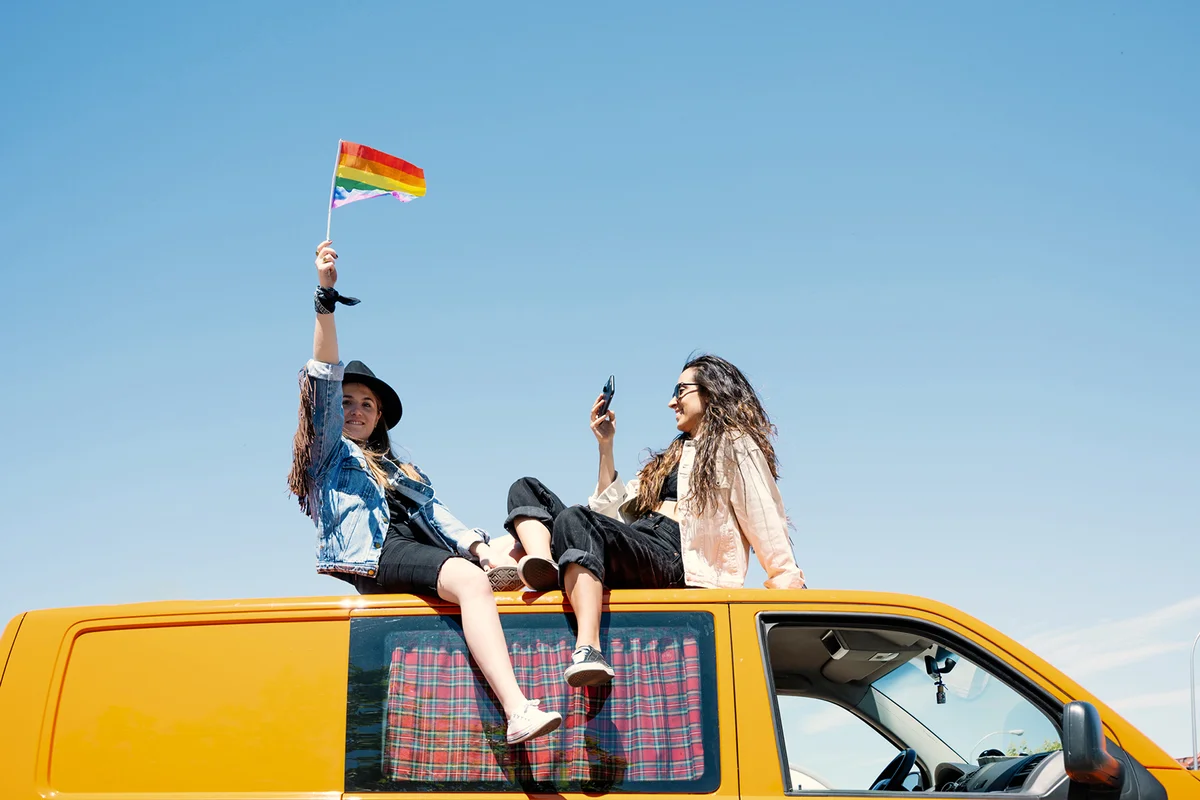 Two women sitting on roof of van, waving pride flag