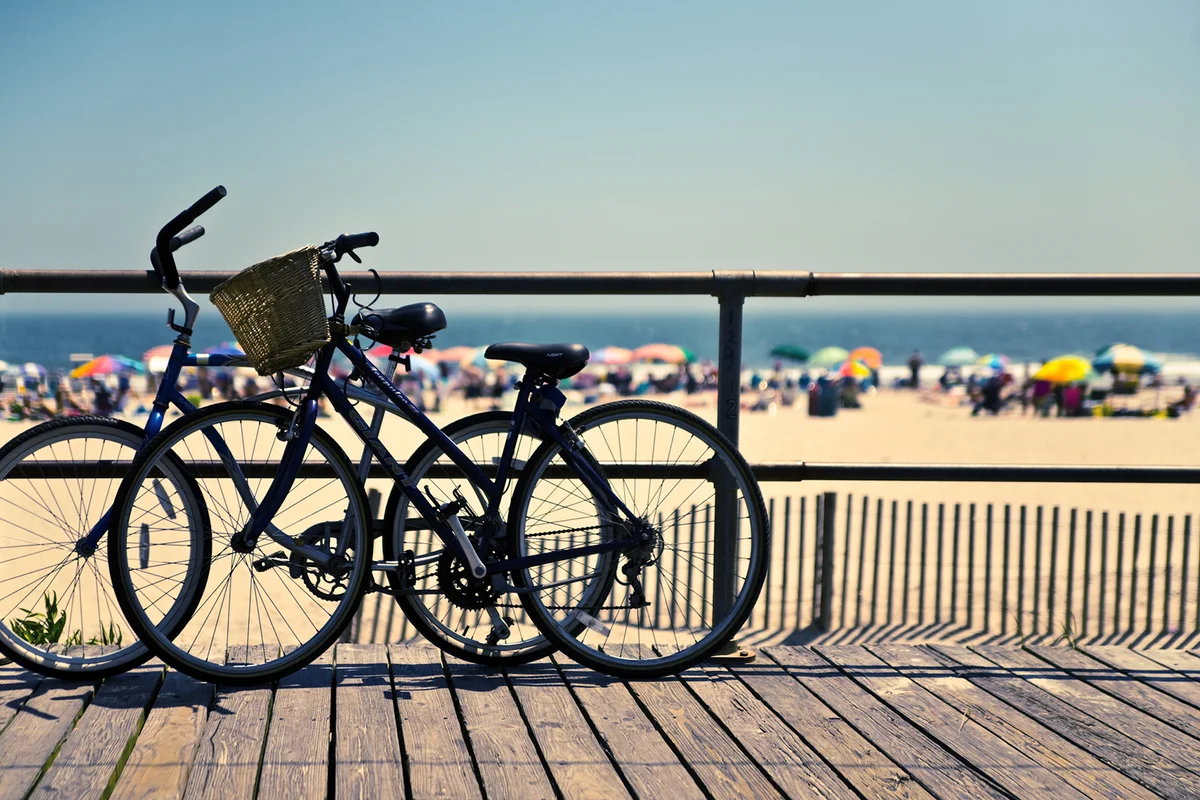 Bicycles in silhouette, propped by the edge of the boardwalk, against a backdrop of a crowded beach in summer.