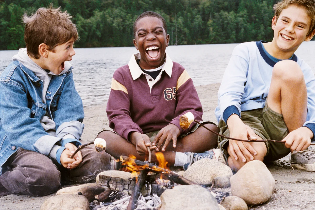 Boys Sit on a Riverbank Toasting Marshmallows Over a Campfire and Laughing