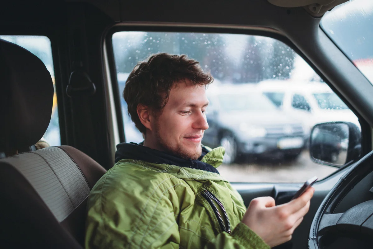 Young smiling man in the green jacket holding smartphone in the car