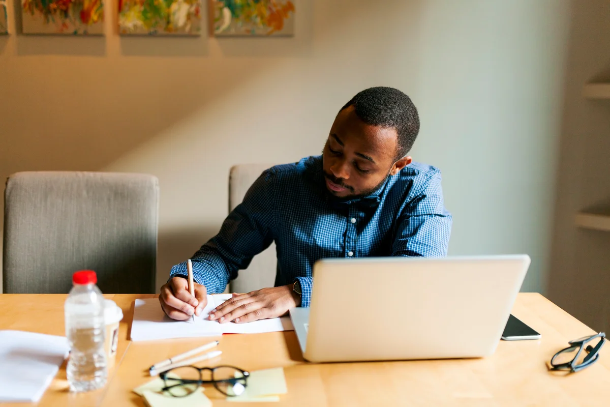 man with laptop writing in notebook