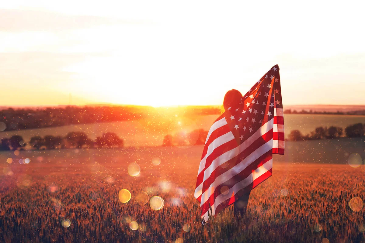 Beautiful young woman with the American flag in a wheat field at sunset with bokeh and sparkle