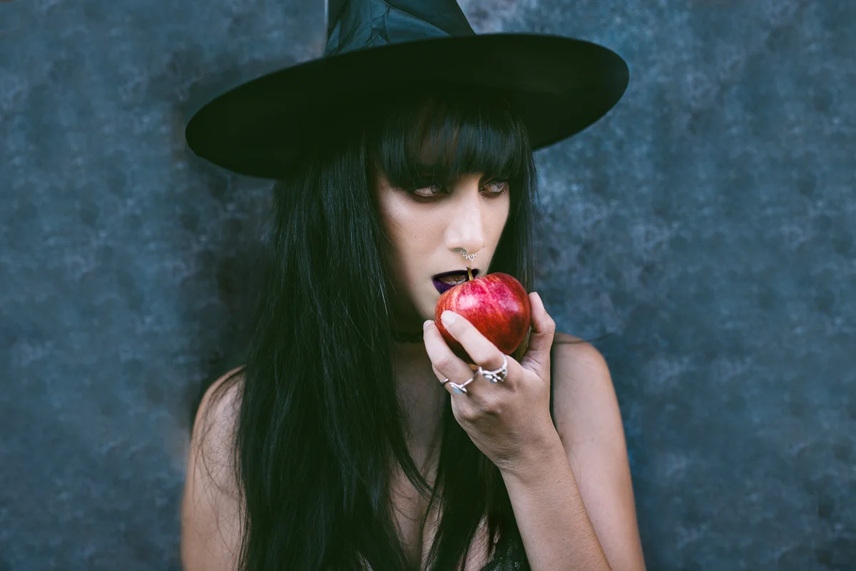Woman Wearing Witch Hat and Makeup While Eating Apple Against Wall