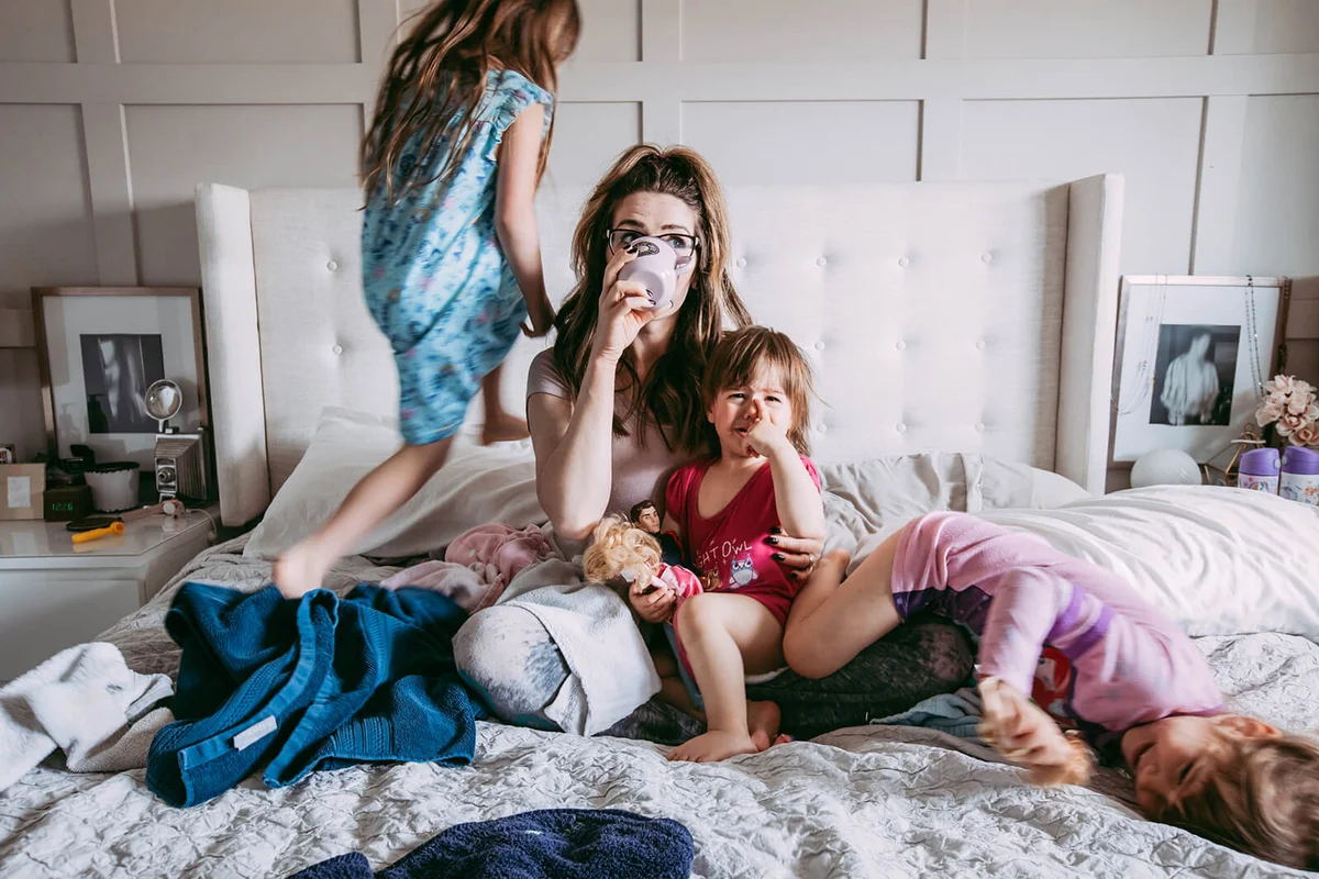 stressed out young mother sipping on a cup of coffee on her messy bed while her three daughters are playing around her