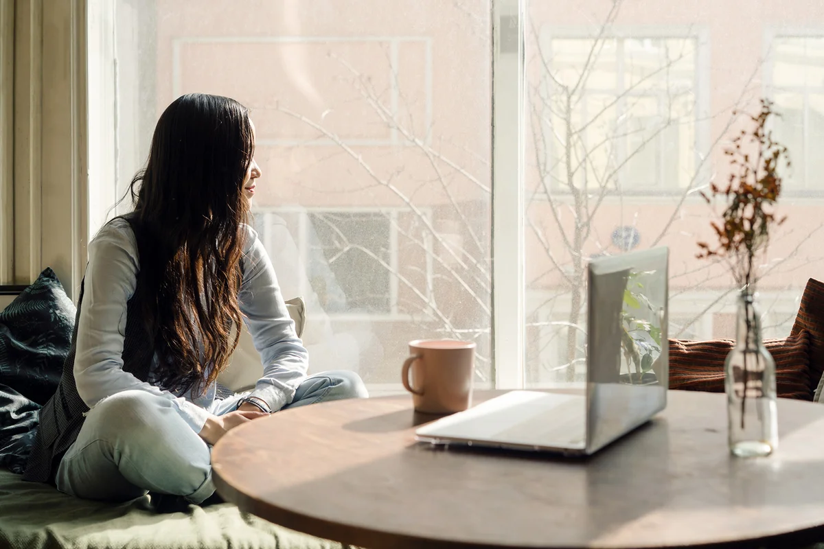 Woman sitting by the window on the sofa with a laptop on a pillow
