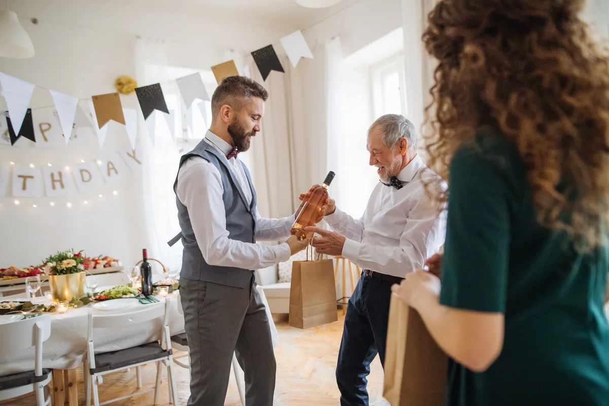 Man Giving A Bottle Of Wine To His Father On Indoor Birthday Party
