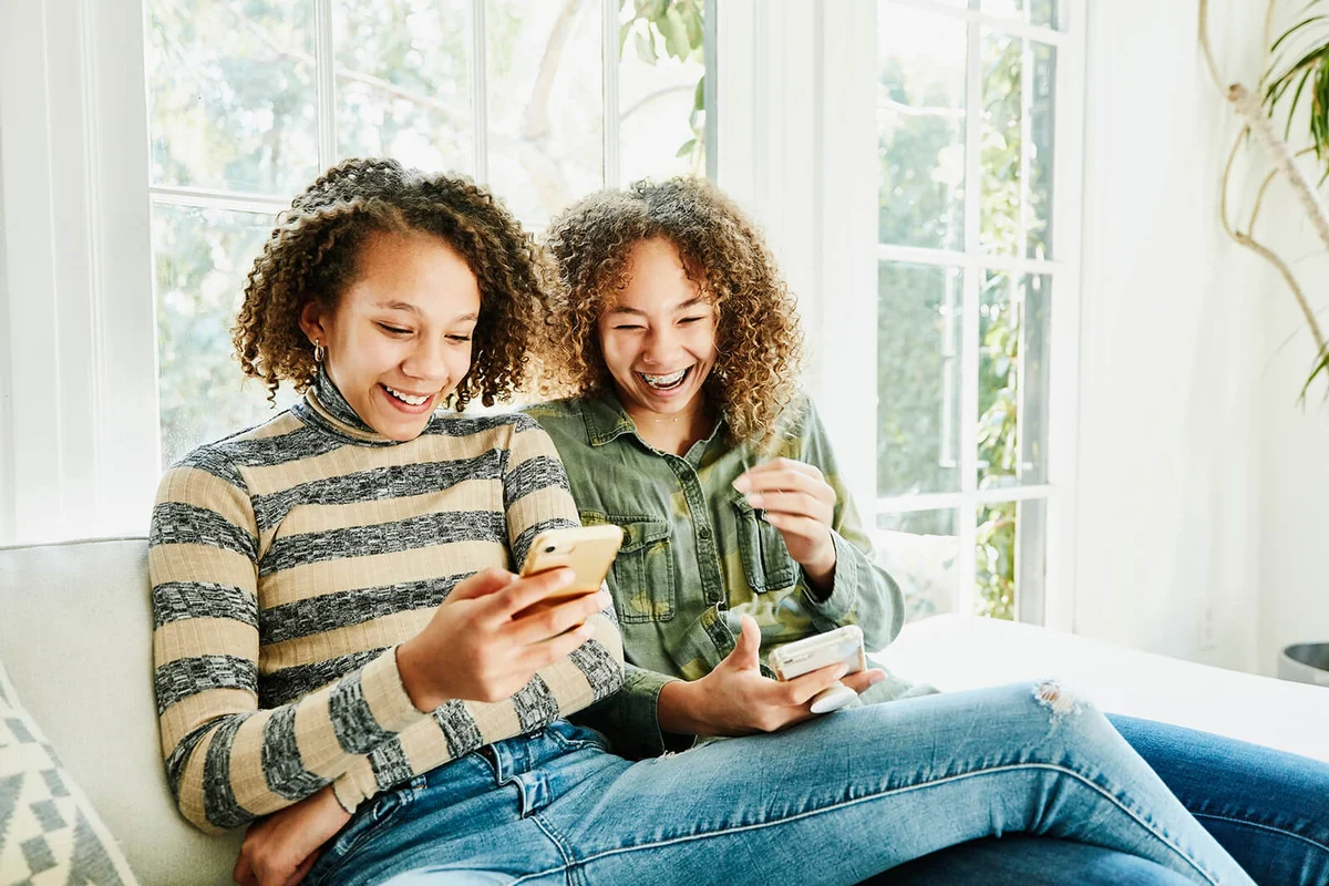 Laughing sisters sitting on couch in living room looking at smart phones