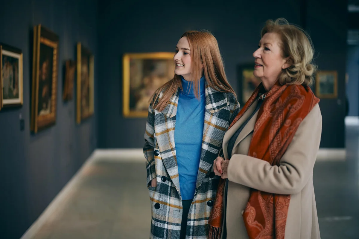 Grandmother and adolescent granddaughter are looking at the paintings in the art gallery