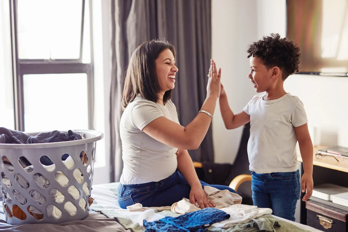 Young mother and young son high fiving while she sits on a bed folding laundry