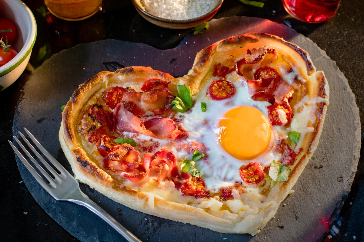 Heart shaped pizza with a fried egg topping on a table surrounded by fresh ingredients and a glass of wine.
