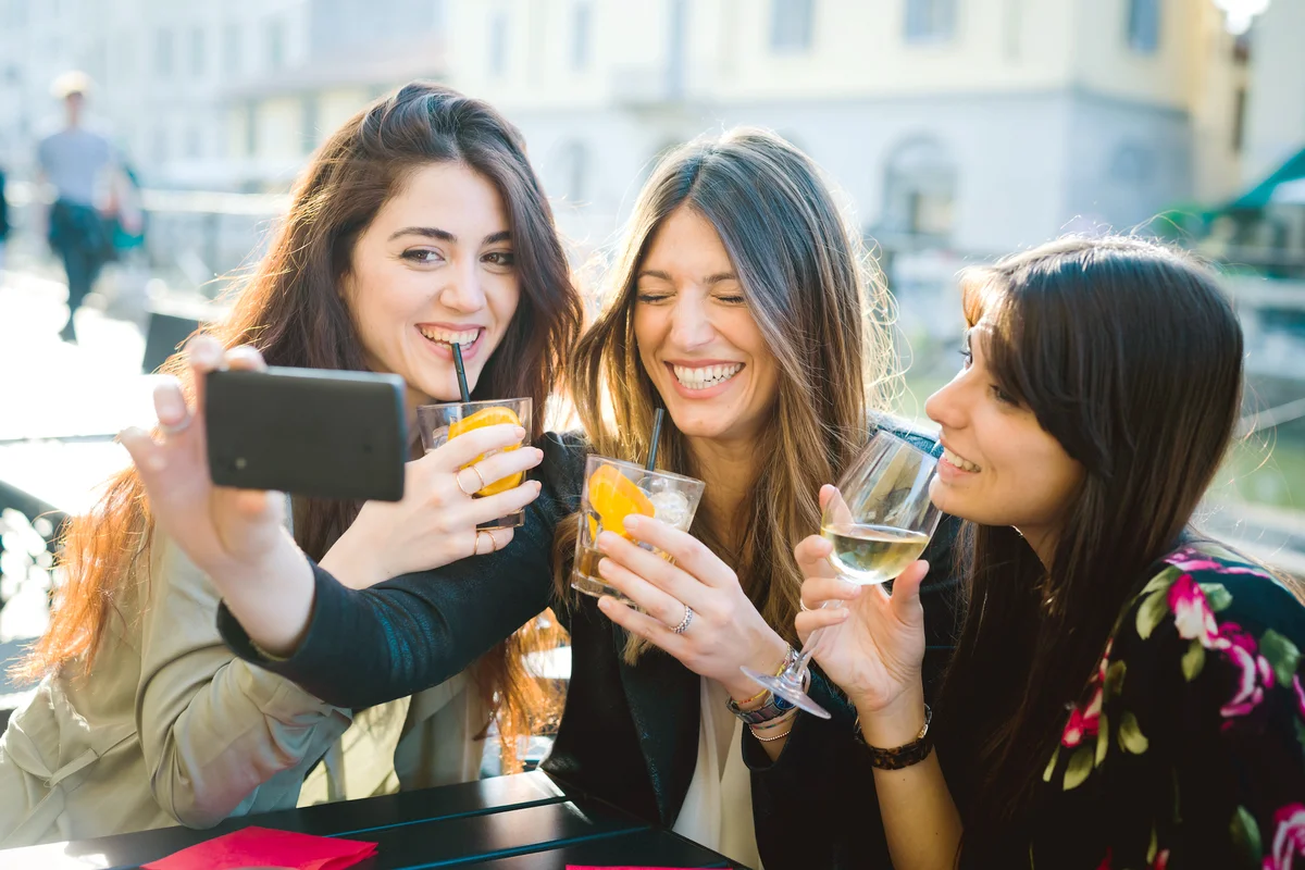 Three young women giggling for smartphone selfie while drinking