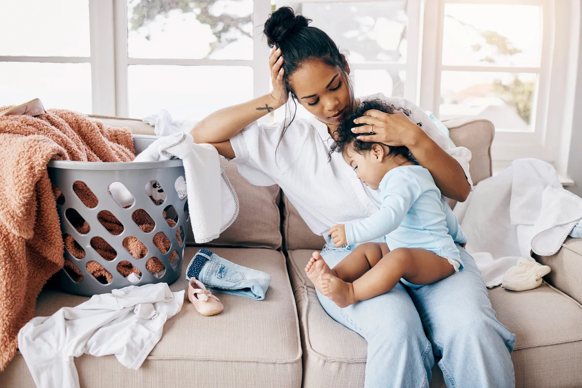 Woman sitting on the sofa comforting her baby daughter while she has laundry to fold