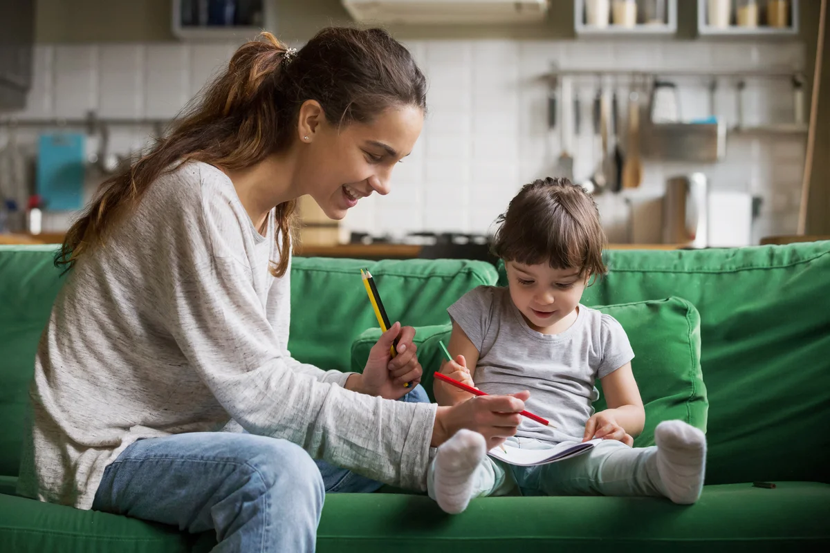 Smiling babysitter and kid drawing while sitting on sofa together