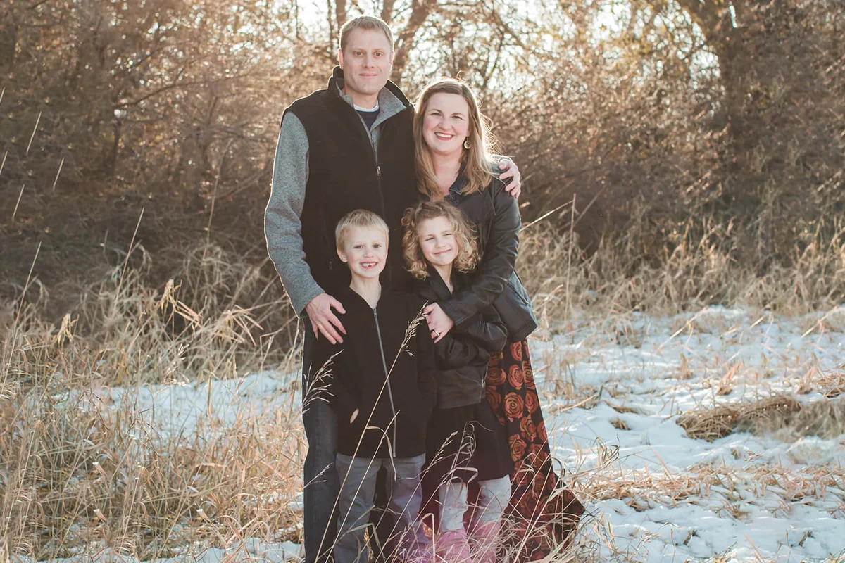 Family of four stand outside on a trail lined with tall pine trees