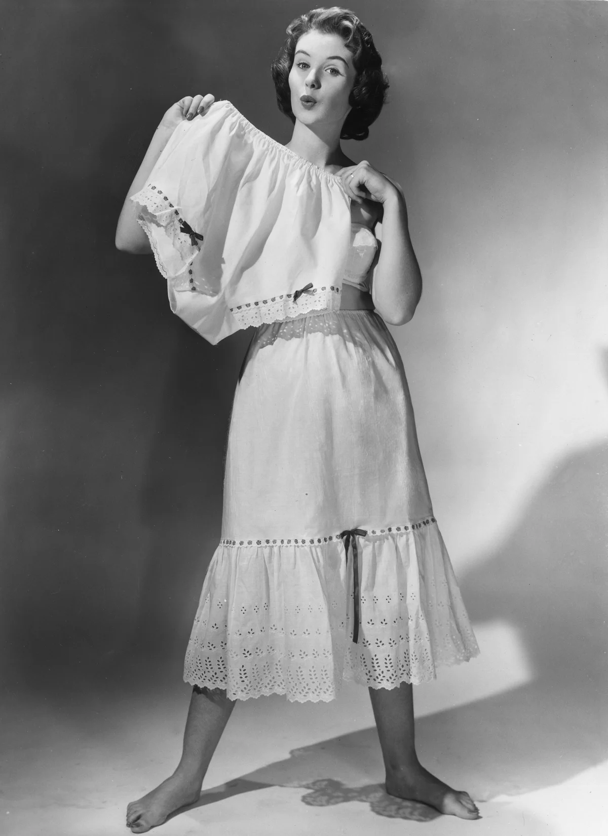 A woman modelling a lace petticoat with a ribbon decoration holds up a pair of matching knickers.