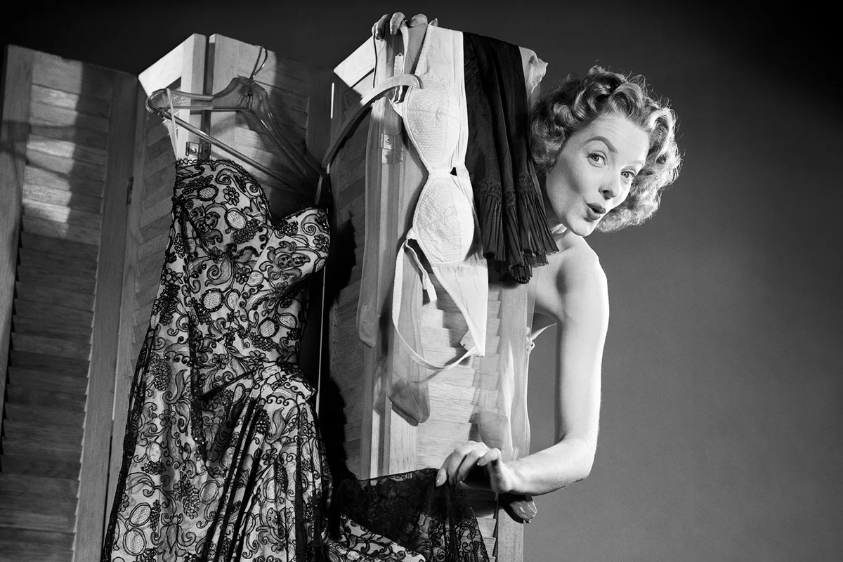 1950s FLIRTING WOMAN LOOKING AT CAMERA PEEKING AROUND FROM BEHIND DRESSING SCREEN HUNG WITH DRESS BRA AND SLIP