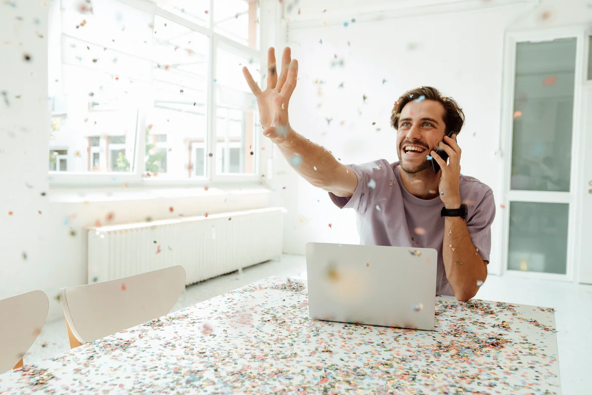 Cheerful man with laptop talking on mobile phone while catching confetti