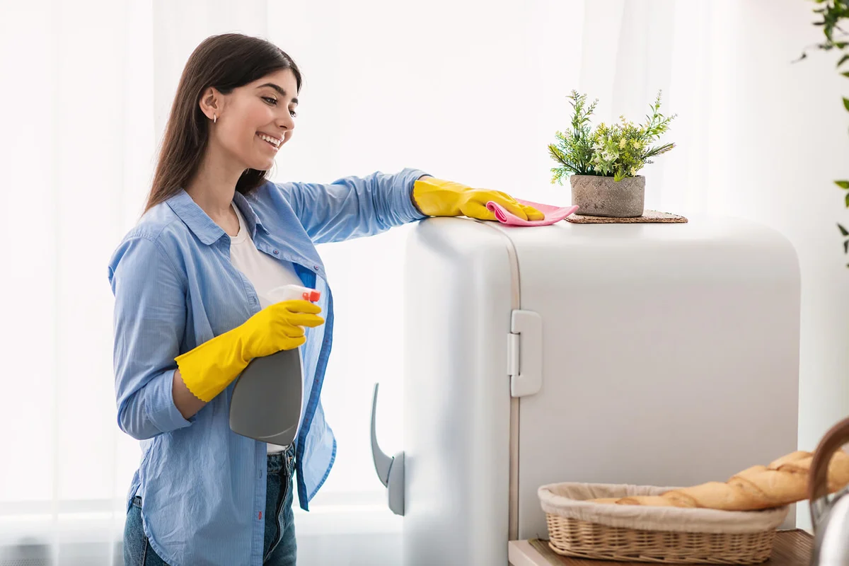 Cheerful young housewife cleaning fridge at kitchen